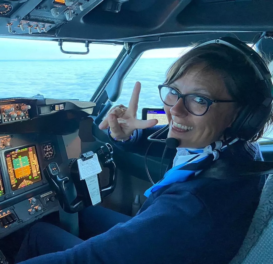 A smiling female pilot wearing a headset and glasses sits in the cockpit of an airplane, making a peace sign with her fingers. She is dressed in a navy blue uniform with a blue scarf, and the aircraft's controls and screens are visible.