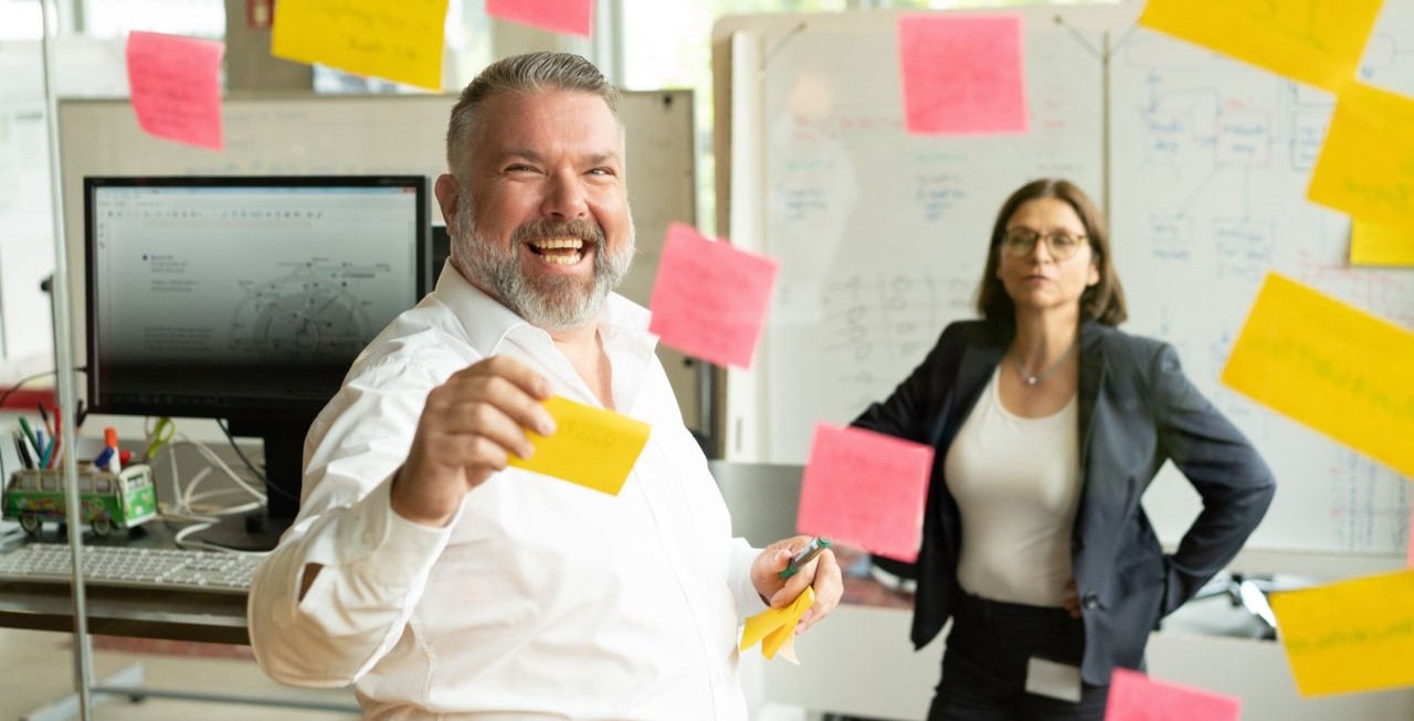 Man working with sticky notes on a screen, woman in the background.