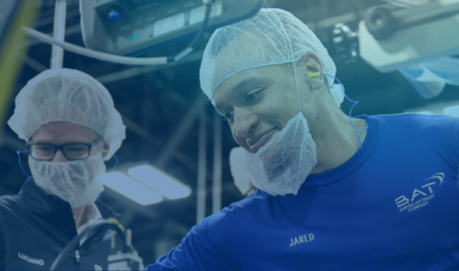Two individuals in hairnets and ear protection in an industrial setting, with visible equipment and branded shirts labeled 'JARED' and 'LUCIANO'