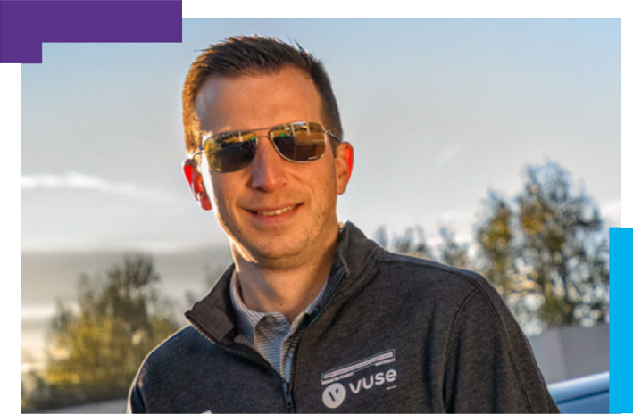 Person wearing a dark gray jacket with the 'Vuse' logo, standing outdoors with trees and a clear sky in the background