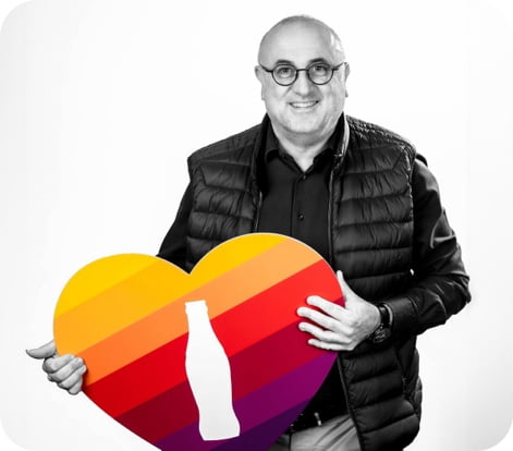 Un homme souriant, en noir et blanc, tient un gros cœur multicolore avec au milieu la forme blanche d’une bouteille de Coca.