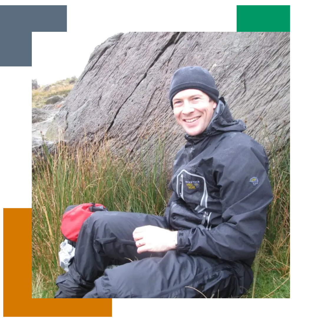 Man sat on top of a mountain in climbing clothes, smiling at the camera