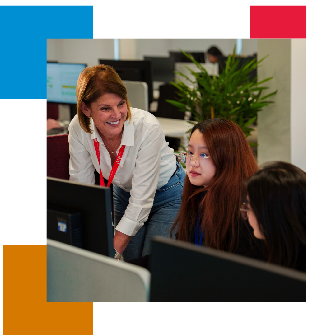 Woman in white shirt and jeans in an office leaning over another woman's desk, laughing and talking whilst working