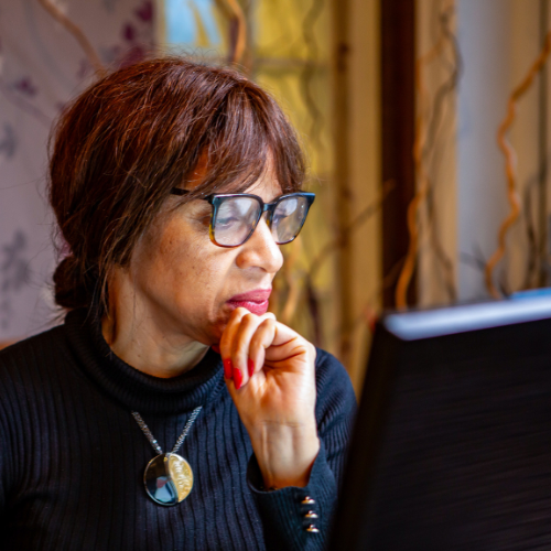 Woman in black top with gold necklace and glasses, working at her laptop screen, looking pensive