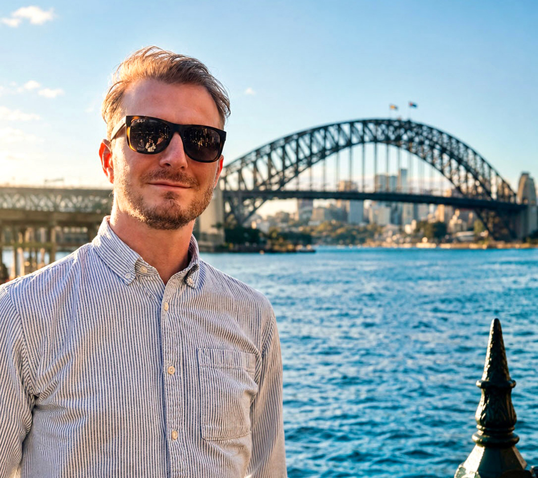 Matthieu is wearing sunglassees and standing in front of a bridge in Sidney