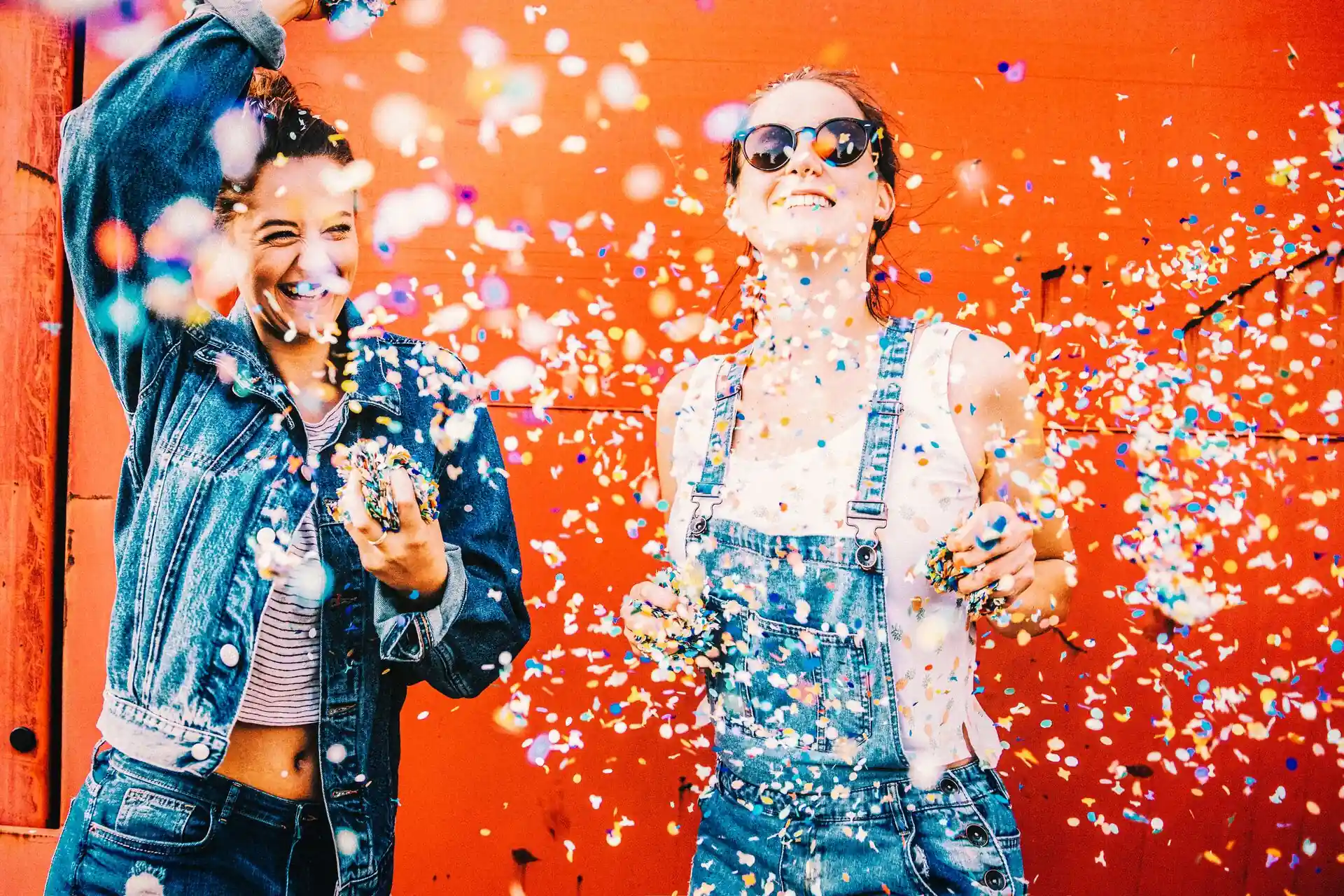 People tossing colourful confetti in front of a bright orange wall, wearing denim clothing, with confetti filling the foreground.