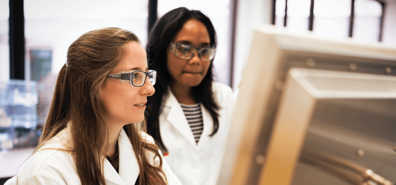 Two women in white lab coats look at a computer screen together