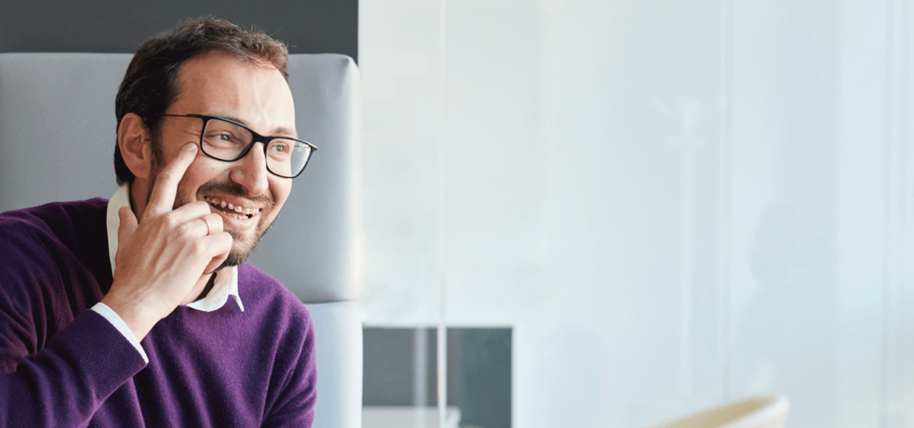 Un homme souriant avec des lunettes, assis au bureau, montre son œil