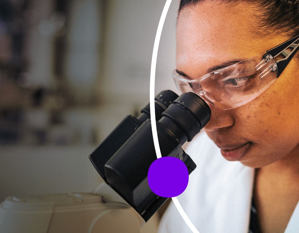 Employee in lab coat and safety glasses looking through microscope