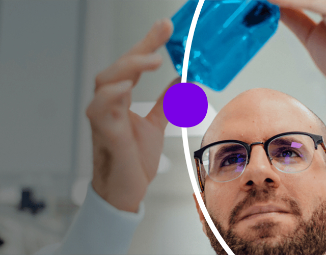 Bearded man in lab, holding up blue specimin jar and looking at it.