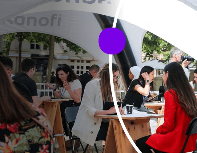 Many groups of people sitting outside, under a tent, at small cafe tables