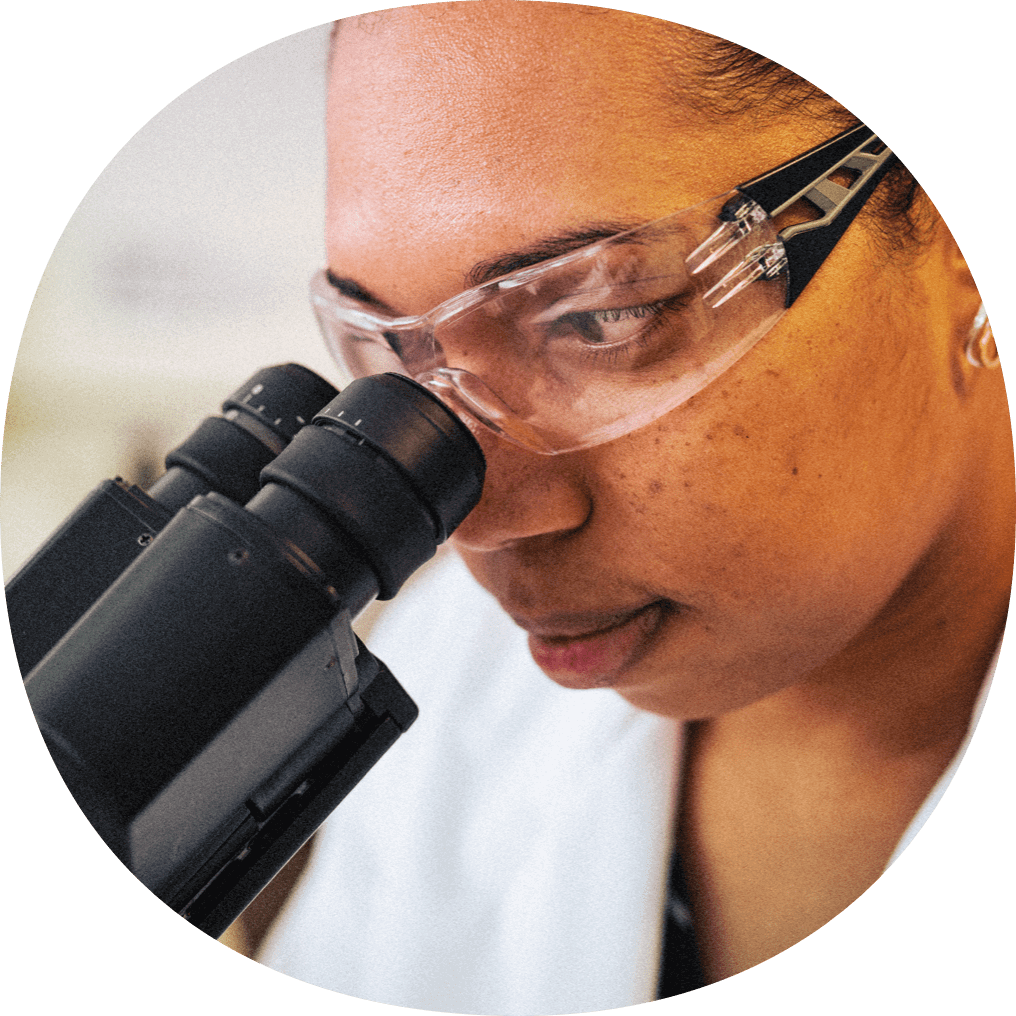 Woman in lab coat and safety glasses looking into a microscope