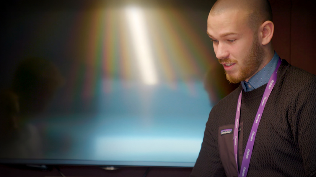 Person wearing a lanyard seated near a large screen in an office.