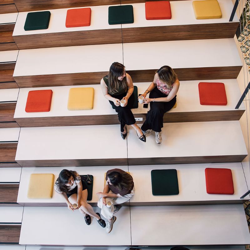 People sitting on tiered wooden steps with colorful cushions, talking and holding coffee.