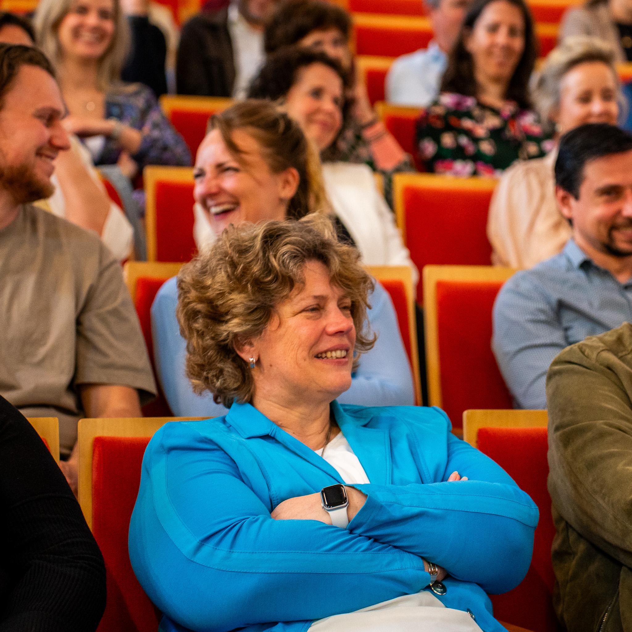 Kenonz collega lachend en zittend in een aula met andere lachende mensen op de achtergrond.