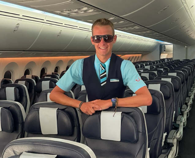 A TUI Cabin Crew male colleague in TUI uniform and sunglasses stands smiling in an empty TUI aeroplane cabin.