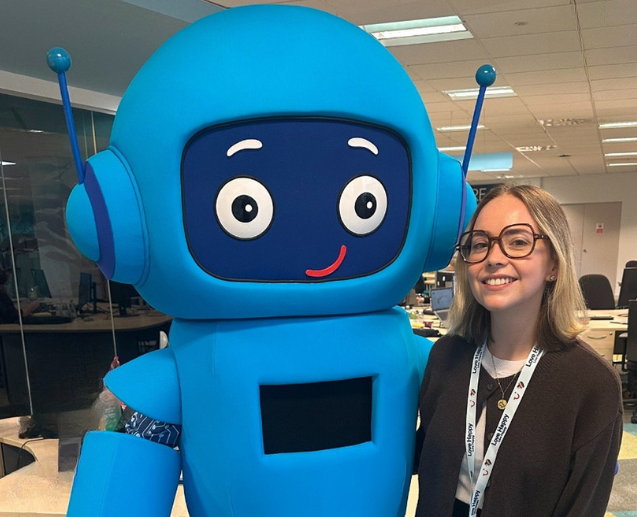 A woman smiles next to a large, cheerful blue robot mascot in an office.