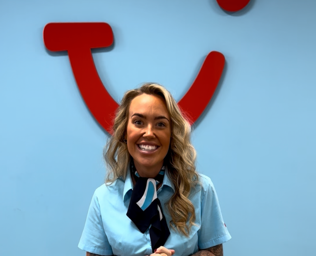 A smiling woman in a TUI Retail uniform and scarf stands in front of a TUI red smile logo on a blue wall.