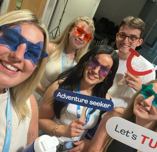 A group of 5 colleagues stand smiling holding props that say 'Let's TUI it' and 'Adventure Seeker'.