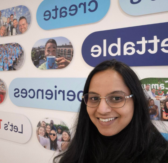 Selfie of a smiling woman with long hair and glasses in front of a wall with photos and motivational words like "Create" and "Experiences" in blue ovals.