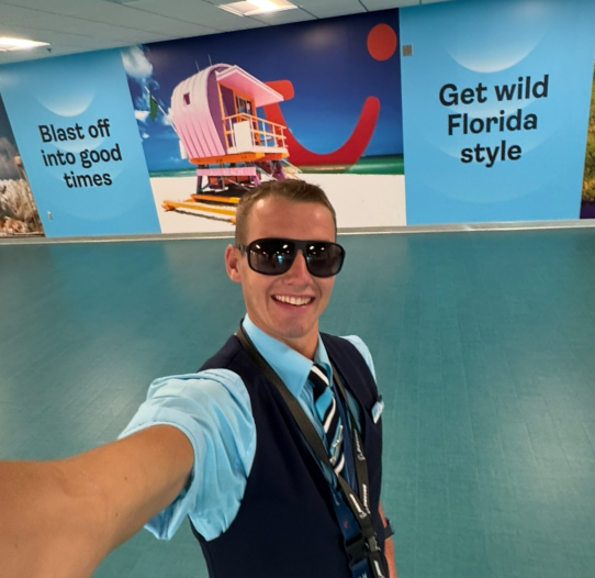 A male colleague in TUI Cabin Crew uniform smiling taking a selfie in front of a TUI sign in Florida.