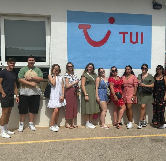 A group of 10 male and female colleagues stood smiling outside the TUI Contact Centre with a blue TUI sign behind them on the wall.