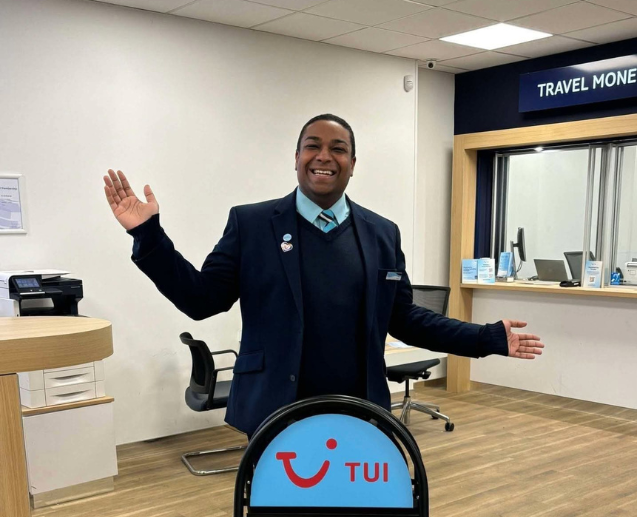A male colleague in TUI Retail uniform stood smiling in a retail store with a blue TUI sign in front of him.