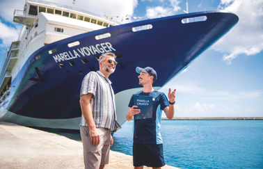 Two men, one in casual clothing and one in Marella Cruises uniform, stand on a sunny dock with the Marella Voyager cruise ship looming large in the background, enjoying a conversation.