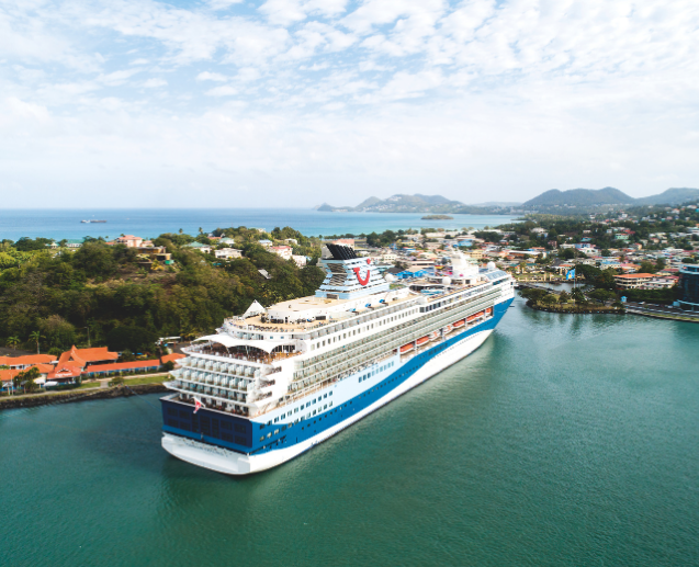A Marella Cruise ship navigates a calm bay, surrounded by vibrant coastal homes and lush hills under a partly cloudy sky.