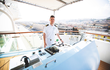 A ship officer in a white uniform stands at the control panel on the bridge of a vessel, with a cityscape and dock visible in the background.