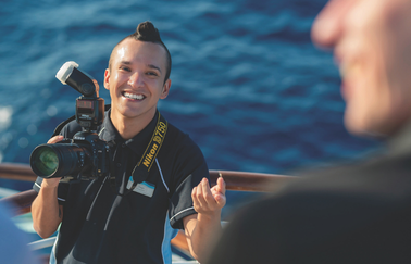 A photographer in uniform on a Marella Cruise ship captures moments against a backdrop of blue ocean, holding a professional camera and smiling. 