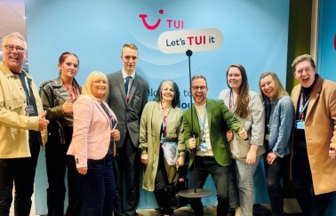 A group of nine people stands smiling in front of a TUI-themed backdrop. They are casually dressed and give a thumbs-up, conveying a cheerful atmosphere.