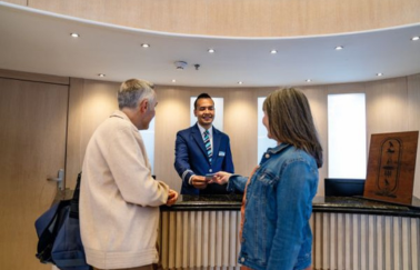 A hotel receptionist in a blue suit smiles as he hands a key card to a middle-aged couple at a modern, warmly-lit reception desk.
