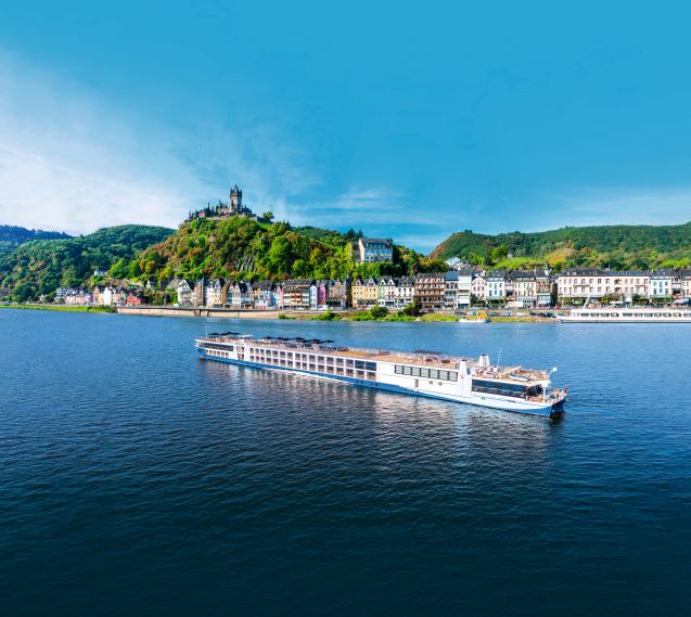 A river cruise ship sails on a wide blue river, with a picturesque town and a hilltop castle in the background under a clear blue sky.