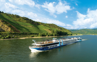 A river cruise ship sails on a calm river bordered by lush, green hills under a partly cloudy sky.