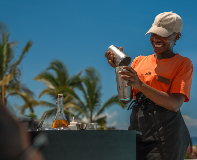 A The Mora colleague stands poolside in front of palm trees and blue skies making cocktails