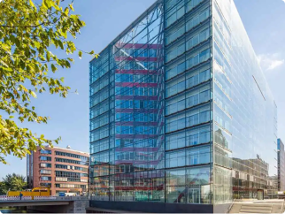 A tall, glass office building surrounded by green trees and a blue sky