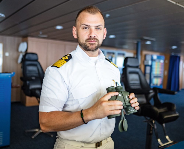 Ship captain in a white uniform holding binoculars stands on a ship's bridge.