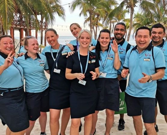 A group of happy people standing together in TUI uniforms on a beach with palm trees, smiling to the camera.