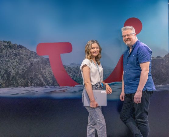 An older man and a younger woman standing in front of a wall with TUI branding on it. They are mid-discussion, looking at the camera only for a moment.