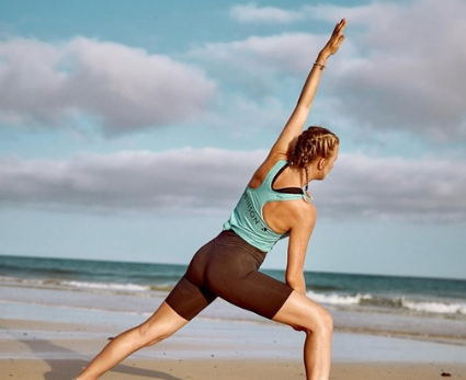 Eine junge Frau in einer Yoga-Pose, im Hintergrund das Meer und ein leicht bewölkter Himmel.