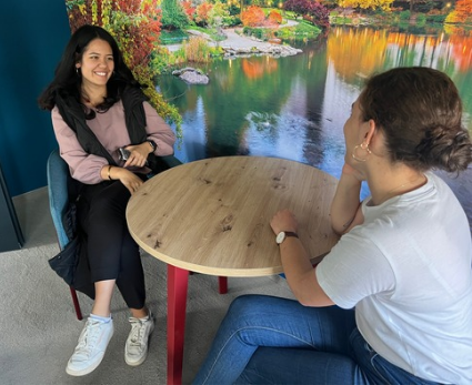 Zwei junge Frauen sitzen entspannt an einem Tisch, im Hintergrund ein großes Bild von einem See in einem Park.