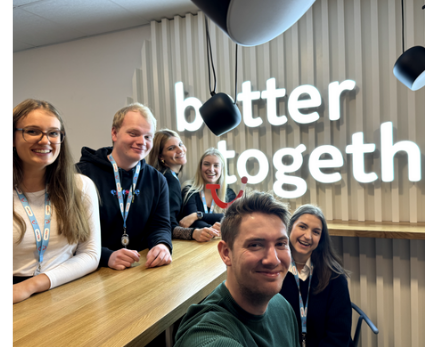 Eine Gruppe von sechs jungen Leuten sitzen vor einer Holz-Wand mit dem LED-Schriftzug "better together"