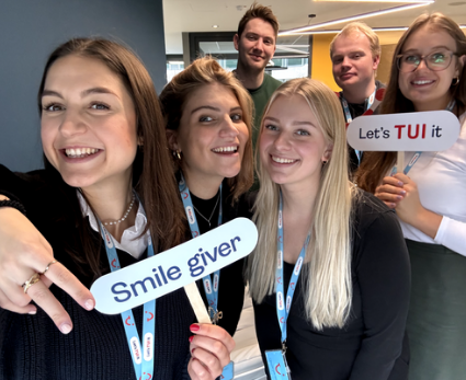 Eine Gruppe von sechs jungen Menschen strahlt in die Kamera. Sie halten zwei Schilder mit den Schriftzügen "Smile Giver" und "Let's TUI it"