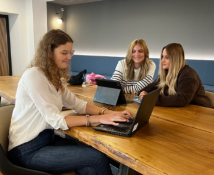 Drei junge Frauen sitzen an einem Holztisch. Eine von Ihnen arbeitet an einem Laptop, die anderen Beiden an einem Tablet.