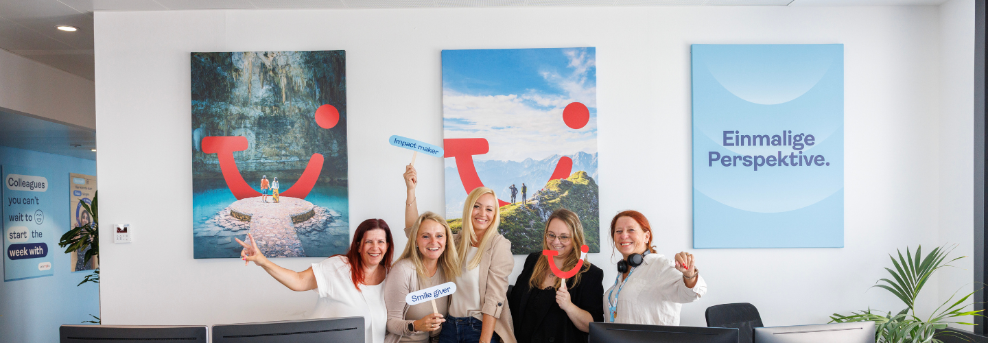 Five smiling women stand together in an office, holding signs "Smile giver" and "#powerofsmile." Three posters with scenic images and German text are on the wall.