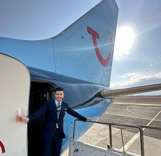 A TUI cabin Crew member in uniform stood at the entrance of a TUI plane smiling.