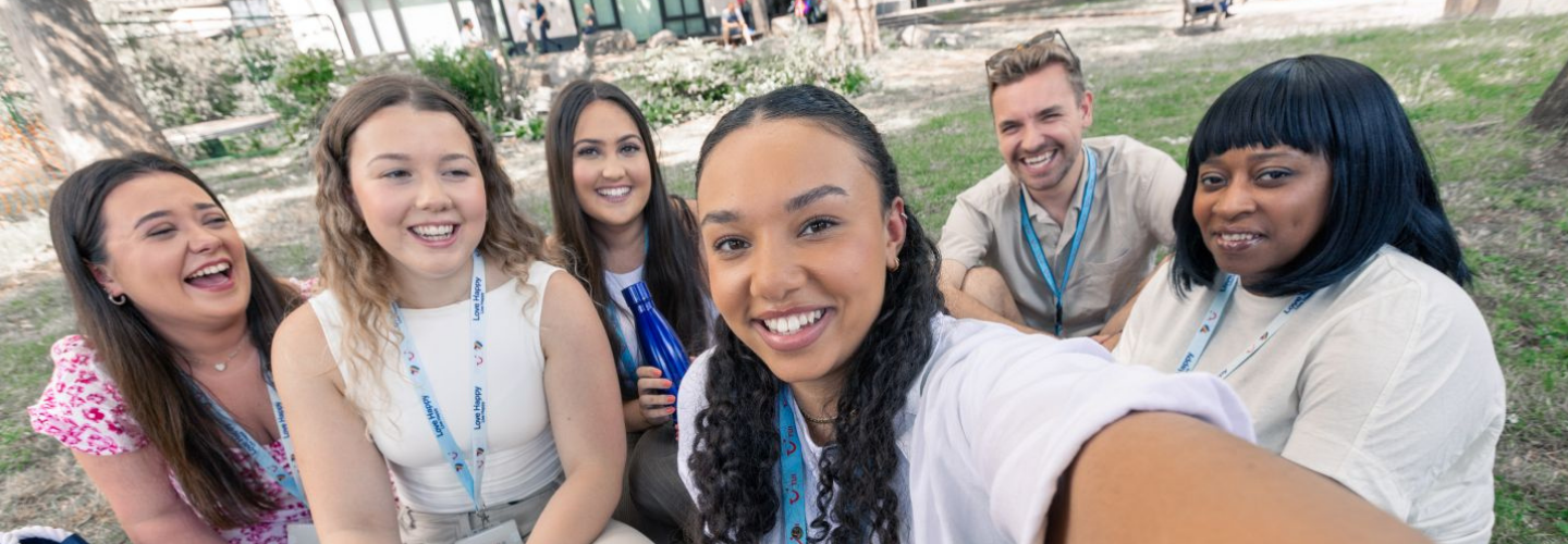 A diverse group of six people sitting on grass, laughing and posing for a selfie. They wear casual attire and lanyards, enjoying a sunny outdoor gathering.
