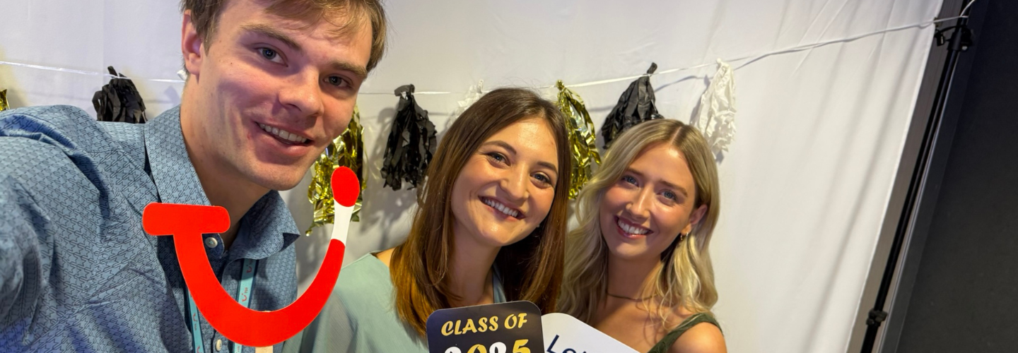Three people smiling at a photo booth with festive black and gold decorations. They hold props, including a "Class of 2025" sign.