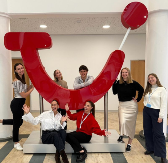 A group of seven people smiling and posing around a large red TUI logo in the office.